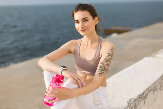 Giovane donna sorridente in top sportivo e leggings bianchi che tiene in mano una bottiglia sportiva rosa mentre guarda sognante in camera con vista mare sullo sfondo