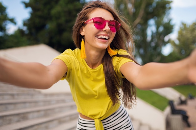 Giovane donna sorridente piuttosto elegante che fa selfie foto nel parco cittadino, positivo, emotivo, con indosso top giallo, occhiali da sole rosa, tendenza moda stile estivo, capelli lunghi, divertirsi