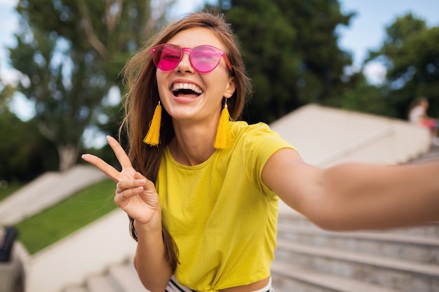 Giovane donna sorridente piuttosto elegante che fa selfie nel parco cittadino, positivo, emotivo, con indosso top giallo, occhiali da sole rosa, tendenza moda stile estivo, capelli lunghi, divertirsi