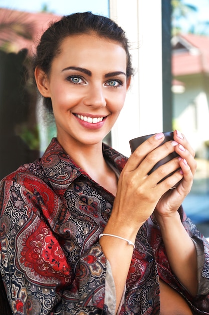 Giovane donna sorridente piuttosto positiva che beve il suo caffè mattutino preferito, ha un bel trucco naturale e una pelle perfetta.
