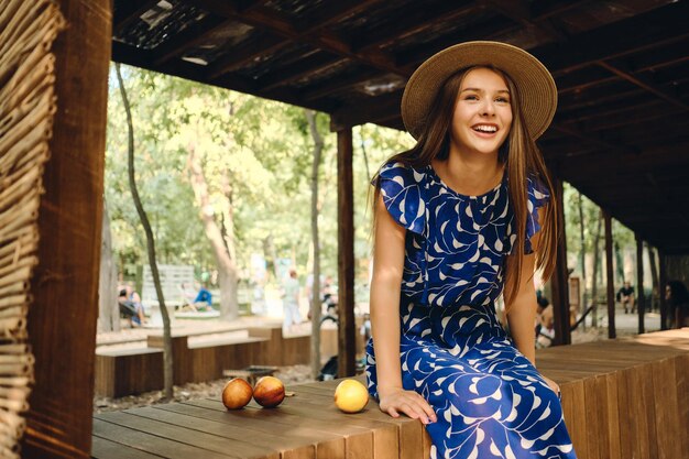 Giovane donna sorridente splendida in abito blu e cappello felicemente seduto con deliziose pesche sulla staccionata di legno nel parco cittadino