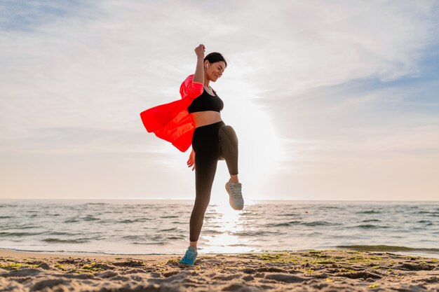 Giovane donna sottile attraente sorridente che fa sport all'alba di mattina che salta sulla spiaggia del mare in abbigliamento sportivo, stile di vita sano, ascolto di musica sugli auricolari, indossa una giacca a vento rosa, divertendosi