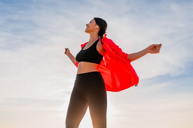 Giovane donna sottile attraente sorridente che fa sport all'alba mattutina ballando sulla spiaggia del mare in abbigliamento sportivo, stile di vita sano, ascoltando musica con gli auricolari, indossando giacca a vento rosa, divertendosi