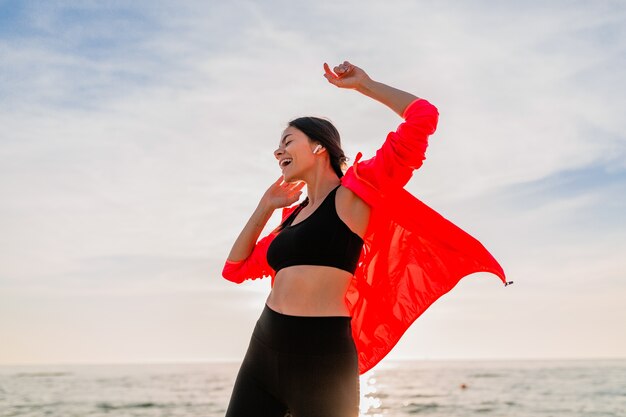 Giovane donna sottile attraente sorridente che fa sport all'alba mattutina ballando sulla spiaggia del mare in abbigliamento sportivo, stile di vita sano, ascoltando musica con gli auricolari, indossando giacca a vento rosa, divertendosi