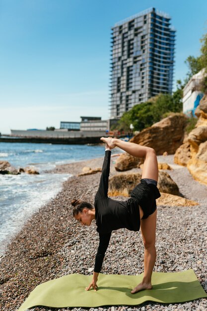 Giovane donna sottile che fa esercizio sulla stuoia di yoga all'aperto alla spiaggia di ciottoli in riva al mare