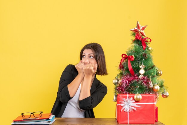 Giovane donna spaventata guardando qualcosa seduto a un tavolo vicino all'albero di Natale decorato in ufficio su giallo