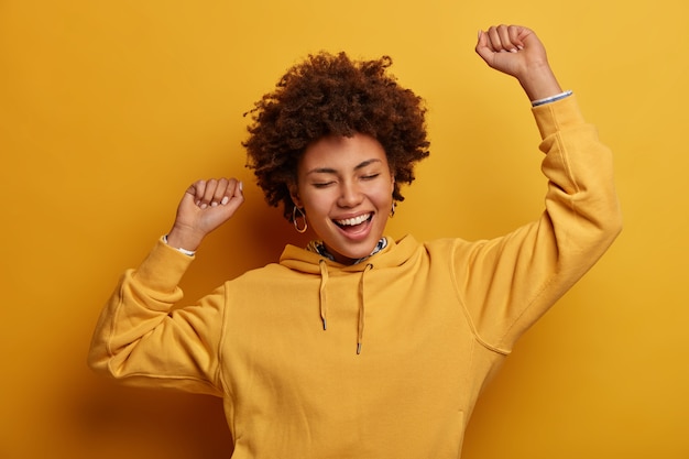 Giovane donna spensierata energica dalla pelle scura balla con le mani alzate, canta la canzone preferita, trionfa sulla vittoria, chiude gli occhi, esprime felicità, ottiene la vittoria o l'approvazione, indossa una felpa gialla