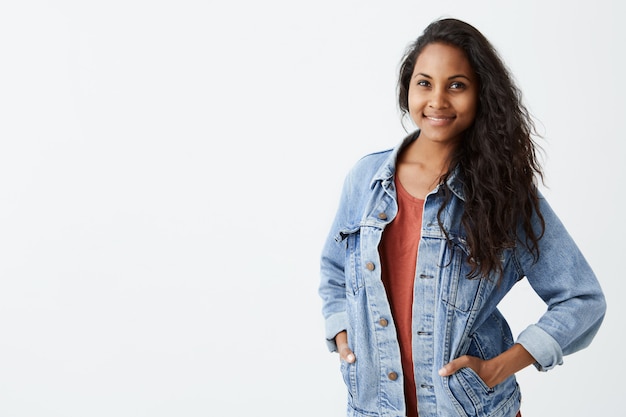 Giovane donna splendida allegra con i capelli ondulati scuri che indossa la giacca di jeans in maglietta rossa che sorride piacevolmente, stando con le sue mani in tasche. Bella ragazza vestita in abiti casuali.