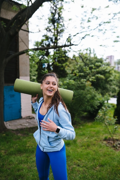 Giovane donna sportiva allegra che cammina nel parco urbano che tiene tappeto fitness.