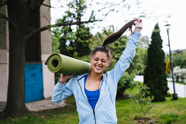 Giovane donna sportiva allegra che cammina nel parco urbano che tiene tappeto fitness.