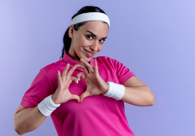 Giovane donna sportiva caucasica felice che indossa la fascia e braccialetti gesti il segno della mano del cuore isolato sullo spazio viola con lo spazio della copia