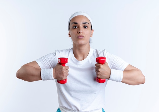 Giovane donna sportiva caucasica fiduciosa che indossa la fascia e braccialetti esercizi con manubri isolati su spazio bianco con spazio di copia