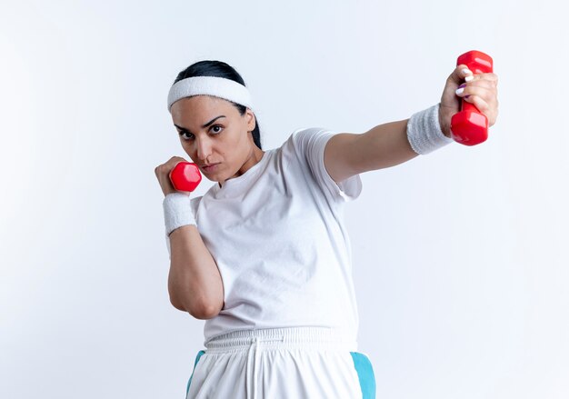 Giovane donna sportiva caucasica fiduciosa che indossa la fascia e braccialetti esercizi con manubri isolati su spazio bianco con spazio di copia