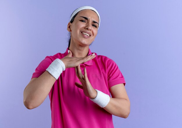 Giovane donna sportiva caucasica infastidita che indossa la fascia e i braccialetti gesti il segno della mano fuori tempo sulla porpora con lo spazio della copia