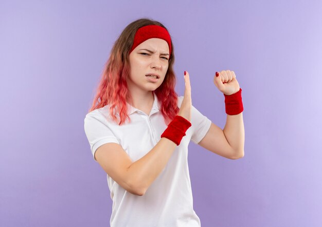 Giovane donna sportiva che fa gesto di difesa con il braccio che stringe il pugno con espressione disgustata in piedi sopra la parete viola