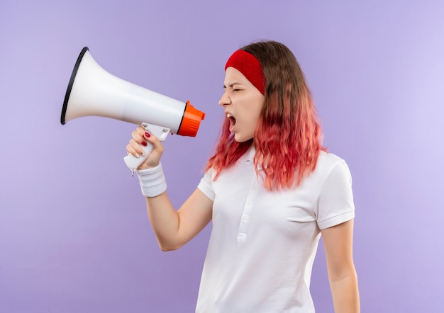 Giovane donna sportiva che grida al megafono con espressione aggressiva in piedi sopra la parete viola