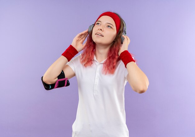 Giovane donna sportiva in archetto con le cuffie che osserva in su godendo la sua musica preferita, formazione con fascia da braccio dello smartphone in piedi sopra la parete viola