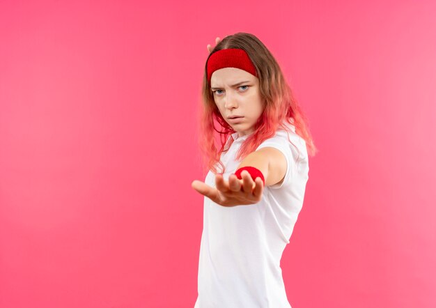 Giovane donna sportiva in fascia che fa il fanale di arresto con la mano con la faccia seria che sta sopra la parete rosa