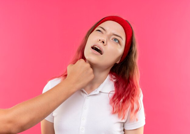 Giovane donna sportiva in fascia ottenendo vomito nella mascella con il pugno in piedi sopra il muro rosa