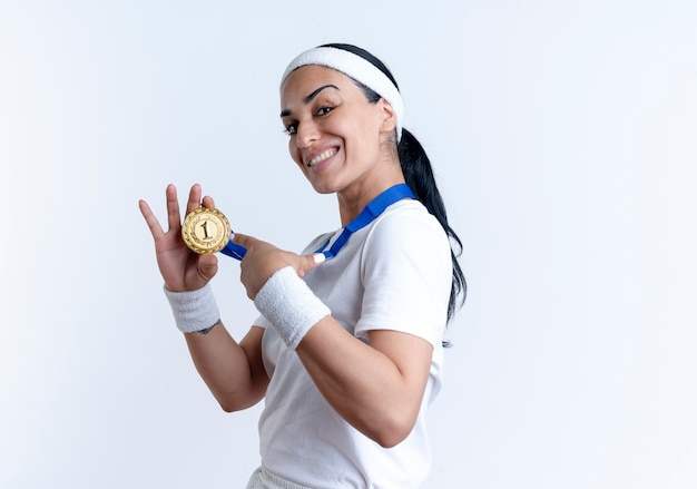 giovane donna sportiva indoeuropea sorridente si trova lateralmente tenendo e indicando la medaglia d'oro