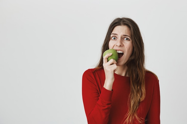 Giovane donna sveglia che morde mela verde e che fa smorfie, mal di denti di sensibilità