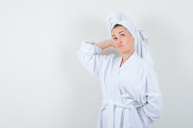 Giovane donna tenendo la mano sulla testa in accappatoio bianco, asciugamano e guardando indeciso, vista frontale.