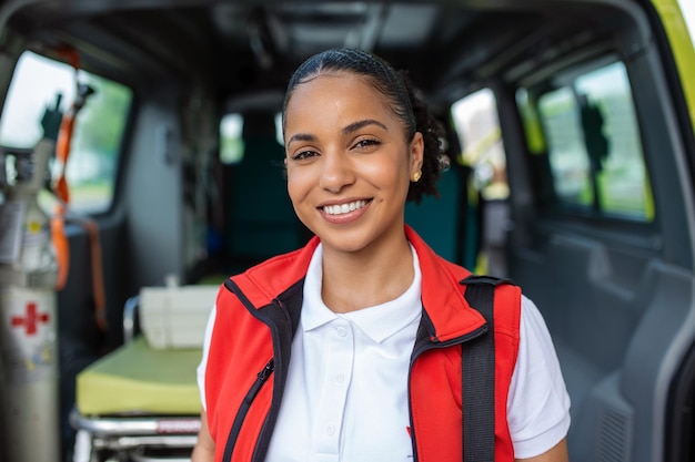 Giovane donna un paramedico in piedi sul retro di un'ambulanza vicino alle porte aperte Sta guardando la telecamera con un'espressione sicura di sé sorridente portando una borsa da trauma medica sulla spalla