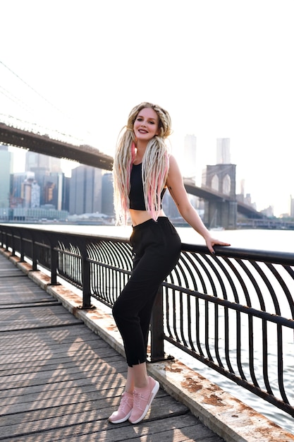 Giovane donna viaggia negli Stati Uniti, posa a New York, splendida vista sul ponte di Brooklyn e sull'isola di manhattan, elegante viaggiatore urbano, insoliti capelli biondi spaventosi, corpo sportivo, viaggio in america.