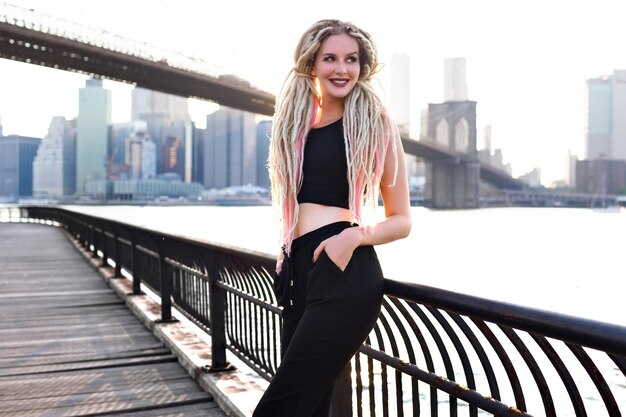 Giovane donna viaggia negli Stati Uniti, posa a New York, splendida vista sul ponte di Brooklyn e sull'isola di manhattan, elegante viaggiatore urbano, insoliti capelli biondi spaventosi, corpo sportivo, viaggio in america.