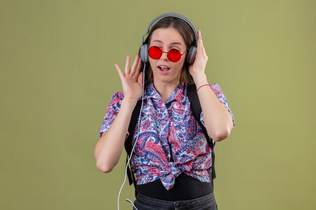 Giovane donna viaggiatore che indossa occhiali da sole rossi e con zaino ascoltando musica utilizzando le cuffie con la faccia felice in piedi su sfondo verde