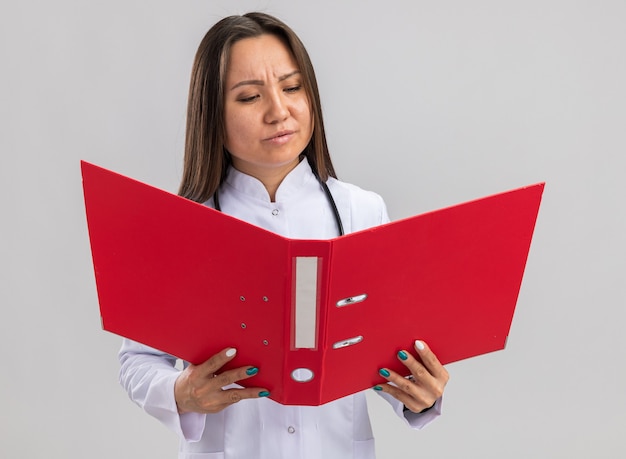 Giovane dottoressa asiatica confusa che indossa tunica medica e stetoscopio che tiene e guarda la cartella aperta isolata sul muro bianco