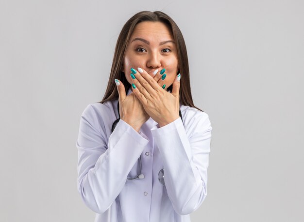 Giovane dottoressa asiatica impressionata che indossa abito medico e stetoscopio guardando la bocca che copre la parte anteriore con le mani isolate sul muro bianco con spazio di copia