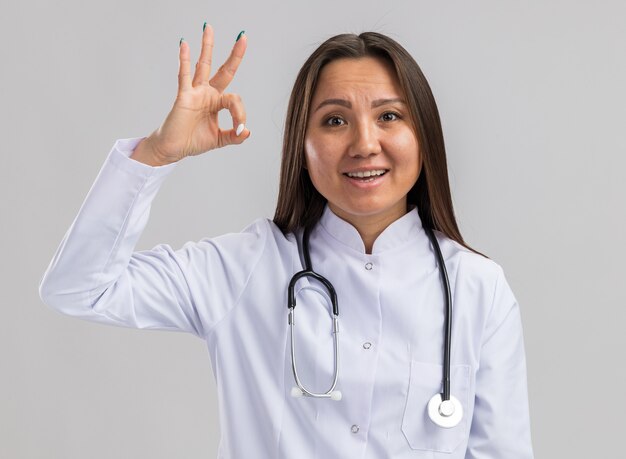 Giovane dottoressa asiatica impressionata che indossa abito medico e stetoscopio guardando la parte anteriore facendo segno ok isolato sul muro bianco