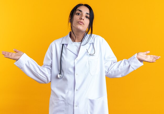 Giovane dottoressa confusa che indossa un abito medico con lo stetoscopio che spande le mani isolate sulla parete gialla