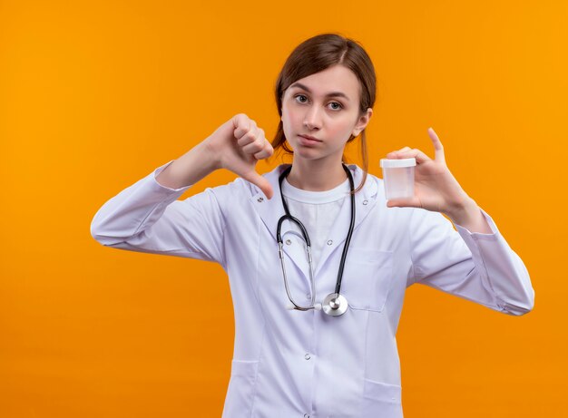 Giovane dottoressa dispiaciuta che indossa abito medico e stetoscopio che tiene il bicchiere e che mostra il pollice verso il basso sulla parete arancione isolata