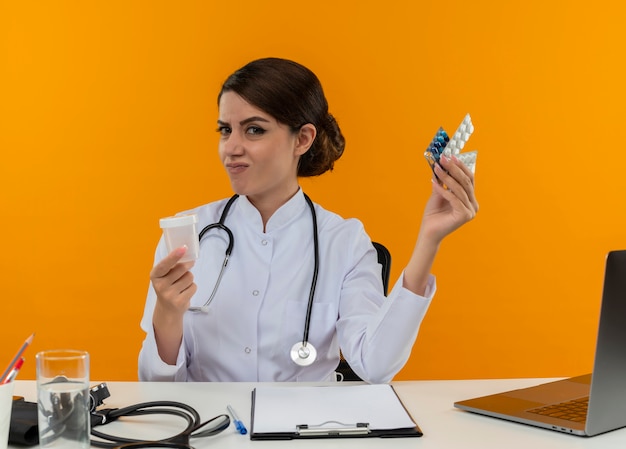 Giovane dottoressa dispiaciuta che indossa veste medica e stetoscopio seduto alla scrivania con strumenti medici e laptop in possesso di farmaci e becher isolato sulla parete gialla
