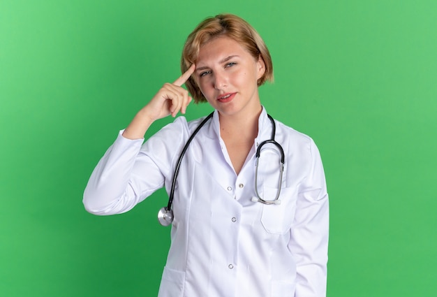 Giovane dottoressa impressionata che indossa un abito medico con uno stetoscopio che mette il dito sulla tempia isolata sulla parete verde green