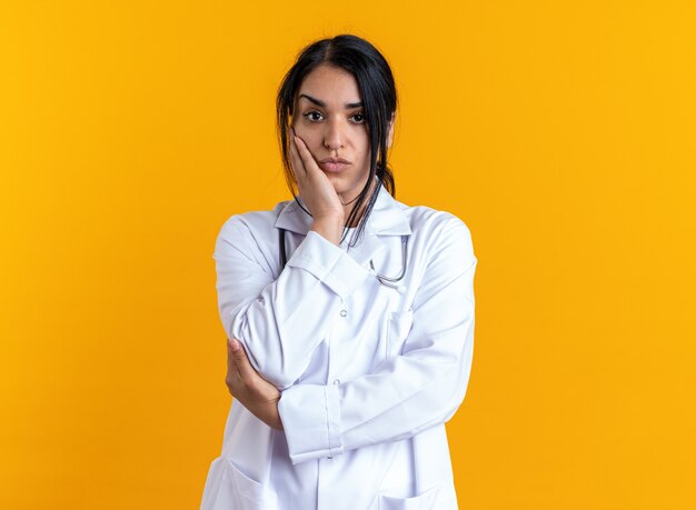 Giovane dottoressa impressionata che indossa un abito medico con uno stetoscopio che mette la mano sulla guancia isolata sul muro giallo