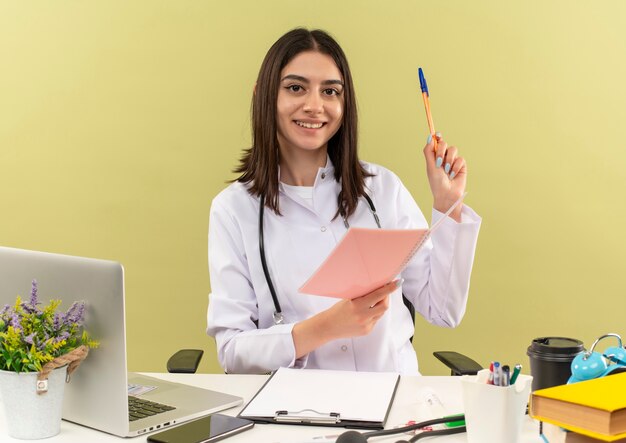 Giovane dottoressa in camice bianco con lo stetoscopio intorno al collo che tiene il taccuino e la penna che guarda al sorriso davanti wirh sul viso seduto al tavolo con il computer portatile sopra la parete chiara