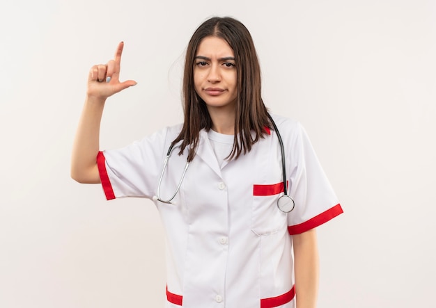 Giovane dottoressa in camice bianco con lo stetoscopio intorno al collo guardando in avanti dispiaciuto mostrando il dito indice in piedi sul muro bianco