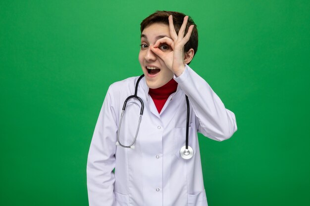 Giovane dottoressa in camice bianco con stetoscopio intorno al collo guardando attraverso le dita facendo segno ok sorridendo allegramente in piedi sul verde