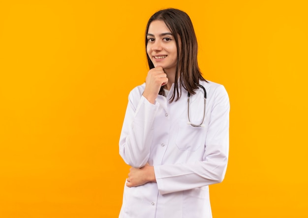 Giovane dottoressa in camice bianco con uno stetoscopio intorno al collo guardando in avanti perplesso in piedi sopra la parete arancione