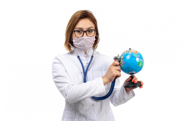 giovane dottoressa in tuta medica bianca con stetoscopio in maschera protettiva bianca che tiene intorno al piccolo globo sul bianco
