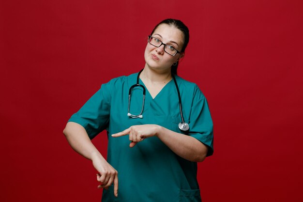 Giovane dottoressa scontenta che indossa occhiali uniformi e stetoscopio intorno al collo guardando la fotocamera che mostra che sei in ritardo gesto isolato su sfondo rosso