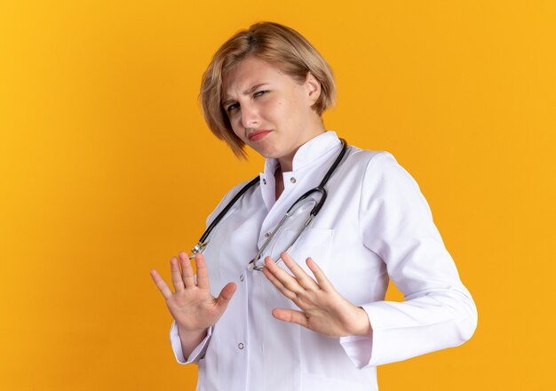 Giovane dottoressa scontenta che indossa un abito medico con uno stetoscopio che mostra il gesto di arresto isolato su sfondo arancione