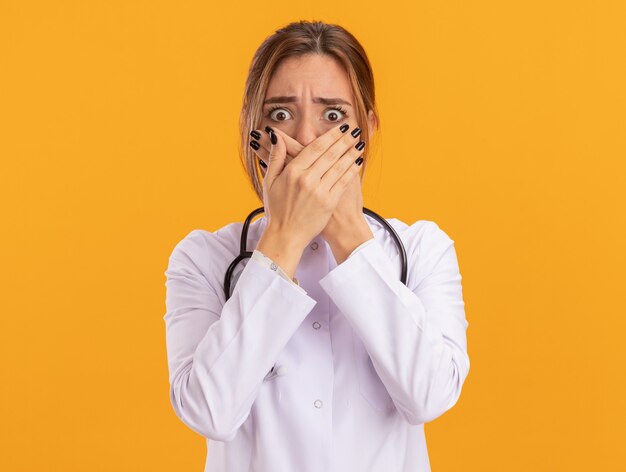 Giovane dottoressa spaventata che indossa un abito medico con uno stetoscopio coperto il viso con le mani isolate sul muro giallo