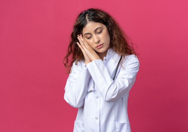 Giovane dottoressa stanca che indossa abito medico e stetoscopio che fa il gesto del sonno con gli occhi chiusi isolati sul muro rosa con spazio per le copie