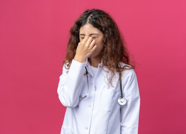 Giovane dottoressa stressata che indossa un abito medico e uno stetoscopio che tiene il naso con gli occhi chiusi isolati sulla parete rosa con spazio per le copie