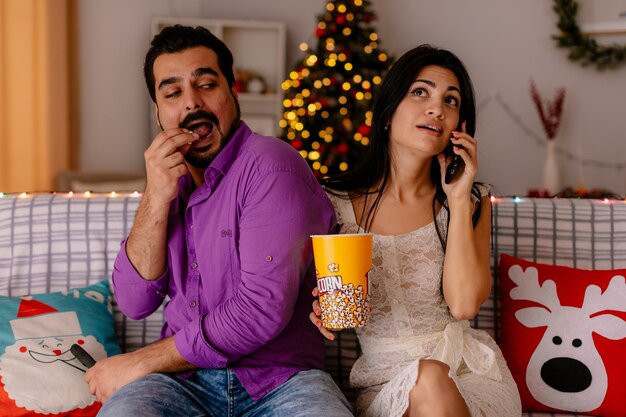 Giovane e bella coppia seduta su un divano uomo mangia popcorn dal secchio mentre la sua ragazza parla al cellulare in camera decorata con albero di natale in background