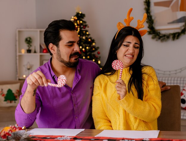 Giovane e bella coppia uomo e donna con bastoncini di zucchero che si divertono insieme felici innamorati nella stanza decorata di natale con l'albero di natale nel muro